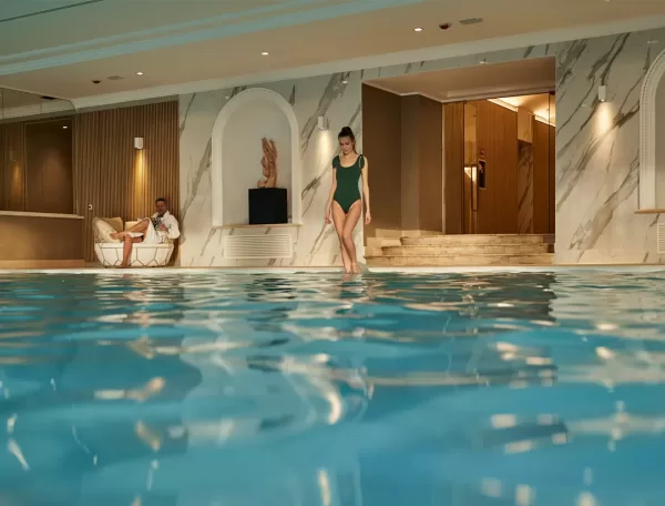 A woman in a green swimsuit walks by an indoor swimming pool, while a man in a white robe relaxes on a lounge chair nearby. The pool area is decorated with marble walls and warm lighting.