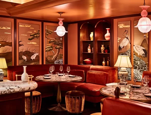Elegant restaurant interior with round marble tables, plush red seating, and decorative screens featuring cranes and trees. Warm lighting from lamps and pendant lights, with shelves displaying vases and ceramics.