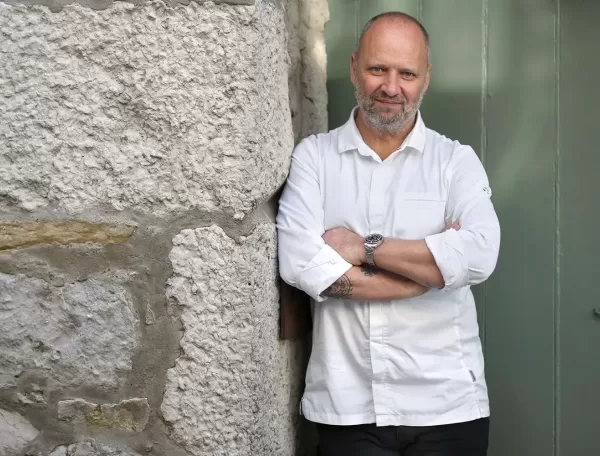 Chef Simon Rogan, wearing a white chefs coat, stands with arms crossed against a stone wall next to a green door, smiling slightly at the camera.