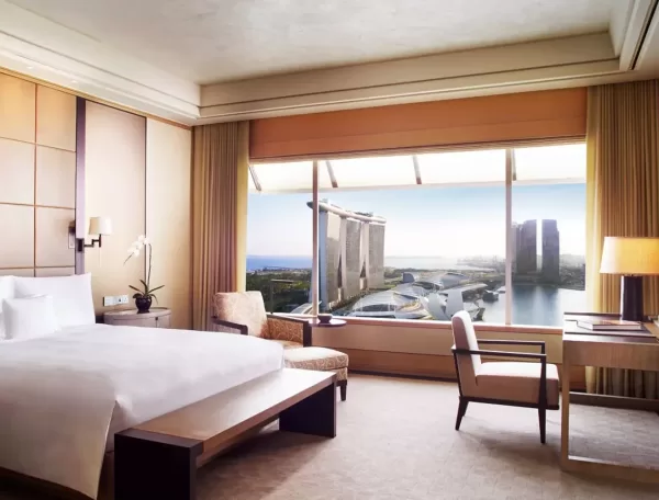 Hotel room with a double bed, armchair, desk, and lamp. Large window offers a view of iconic buildings and a body of water. Neutral tones and wood accents create a cozy atmosphere.