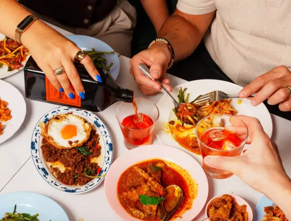Three people share a colorful meal at a table, serving dishes like curry, a fried egg over minced meat, and salad. One person pours a dark drink into a glass while others toast or eat with forks.