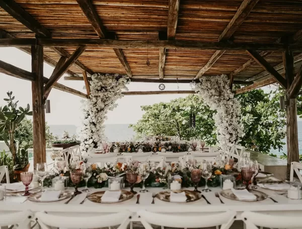 A decorated outdoor dining area under a wooden pergola, featuring long tables set with elegant tableware, floral centerpieces, and white flowers arching around the structure, with greenery and the ocean visible in the background.
