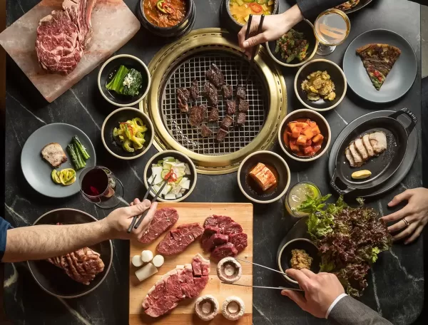 Top view of a Korean BBQ meal, featuring a grill with meat cooking, surrounded by side dishes, raw meats, and hands holding chopsticks and cutting meat. There are plates of vegetables, sauces, and drinks on a dark table.