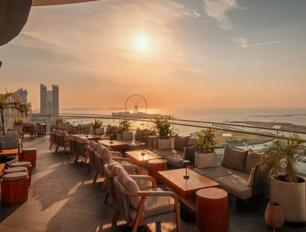 A stylish rooftop terrace with wooden tables and cushioned chairs overlooks the sea at sunset, featuring city buildings and a large Ferris wheel in the distance under a hazy sky.
