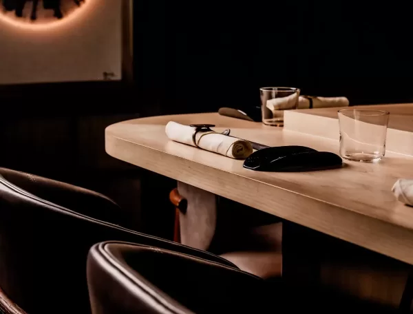 A close-up of a restaurant counter set with empty glasses, rolled napkins, and chopsticks on a light wooden surface, with dark leather chairs and low, warm lighting in the background.