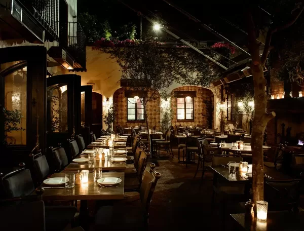 A warmly lit outdoor restaurant patio at night, featuring rustic stone walls, wooden tables set with plates and candles, cushioned chairs, potted plants, and string lights creating a cozy, inviting atmosphere.