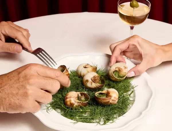 Two hands reach for escargot on a white plate garnished with fresh herbs. A cocktail with an olive sits in the background on a white tablecloth.