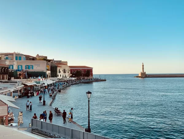 A waterfront promenade with people walking along cafes and shops, colorful buildings to the left, and a lighthouse on a distant pier under a clear blue sky beside calm sea waters.