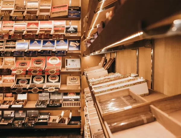 Wooden shelves display various boxes and rows of cigars in a well-lit humidor or cigar shop, with different brands and packaging visible.