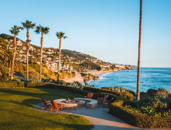 Sunny coastal view with palm trees, a fire pit area surrounded by chairs, manicured grass, and ocean waves. Hills with houses overlook the sandy beach and blue sea under clear skies.