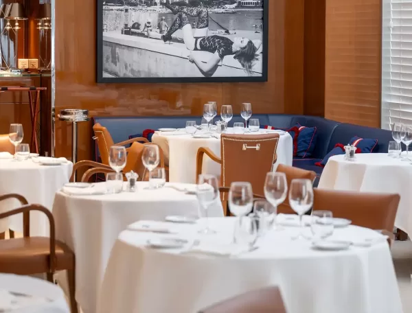Elegant restaurant interior with round tables covered in white tablecloths, set with glassware and cutlery. Brown leather chairs surround the tables. A black and white photo hangs on a wood-paneled wall in the background.