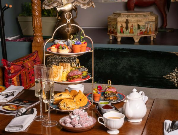 A wooden table with 2 glasses of prosecco, a China cup or tea and a teacup. There is also a 3 tier cake stand with pastries, clotted cream and a mic of coloured cakes, jam and butter,