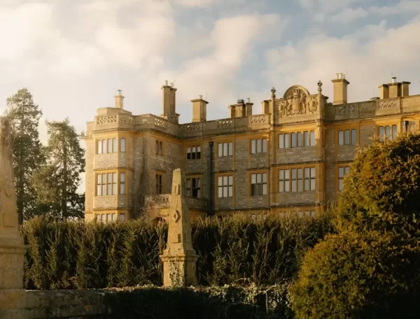 A grand stone mansion with many windows, ornate architectural details, and multiple chimneys stands behind tall hedges on a sunny day, with a lightly clouded sky above.