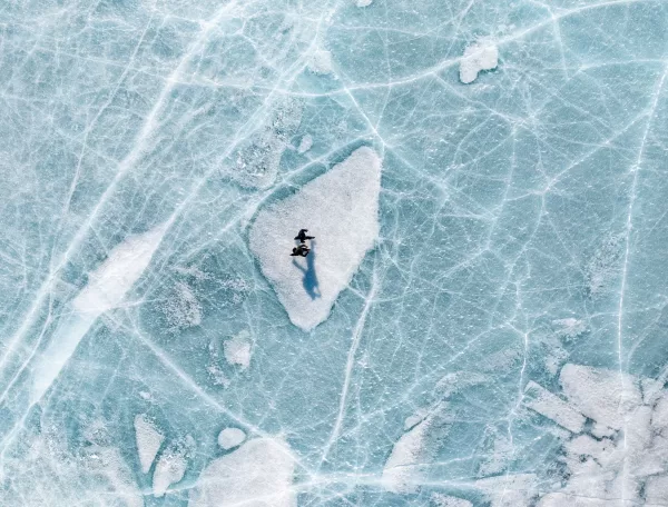 A person stands on a white patch of ice, surrounded by blue cracked and textured frozen lake surface, casting a long shadow. The scene is viewed from above.