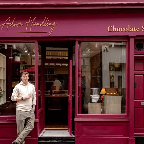 A man in a light sweater and khaki pants stands, holding a drink, outside a maroon storefront that reads Adam Handling Chocolate Shop with large windows displaying chocolates inside.