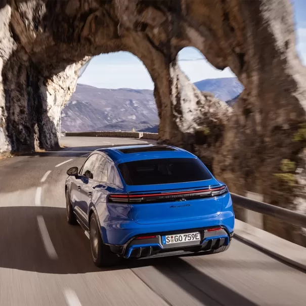 A blue sports car drives along a winding mountain road, passing through a natural rock arch with scenic cliffs and distant hills in the background under a partly cloudy sky.