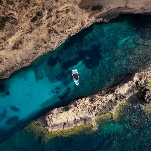 Aerial view of a small boat floating in clear turquoise water between rocky shorelines, with underwater rocks visible and sunlight illuminating the scene.