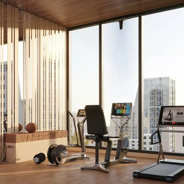 A modern gym room with large windows overlooking tall buildings, featuring a treadmill, two stationary bikes, exercise balls, and wooden decor elements on a wood floor.