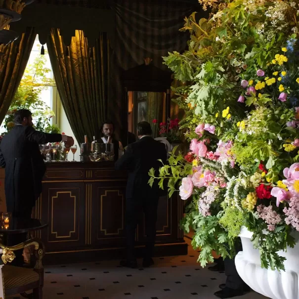 Three people in suits stand at a dark wood bar in an elegant room with heavy drapes. A large, vibrant flower arrangement in a white vase dominates the foreground on the right. Natural light comes in through a window.