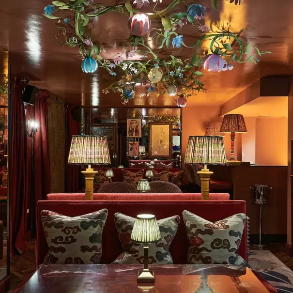 A cozy, dimly lit lounge with red velvet sofas, patterned pillows, and table lamps. Floral light fixtures hang from the ceiling, and mirrors and warm amber lighting create a rich, inviting atmosphere.