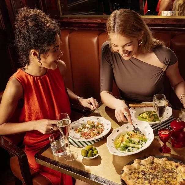 Two women sit at a restaurant table enjoying pasta dishes, salad, and pizza. They are smiling and conversing, with glasses of water and wine in front of them on a cozy brown leather booth.