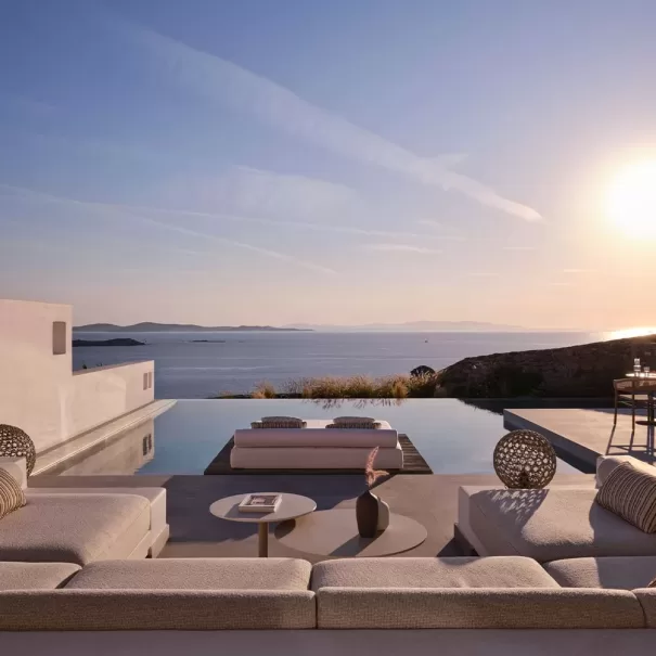 Modern outdoor lounge area with plush seating and a coffee table beside an infinity pool, overlooking the ocean and distant hills at sunset. Warm sunlight bathes the scene, creating a tranquil and luxurious atmosphere.
