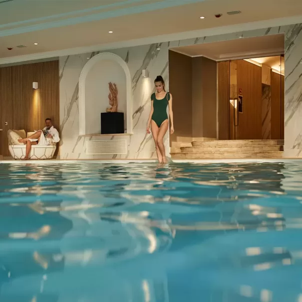 A woman in a green swimsuit walks by an indoor swimming pool, while a man in a white robe relaxes on a lounge chair nearby. The pool area is decorated with marble walls and warm lighting.