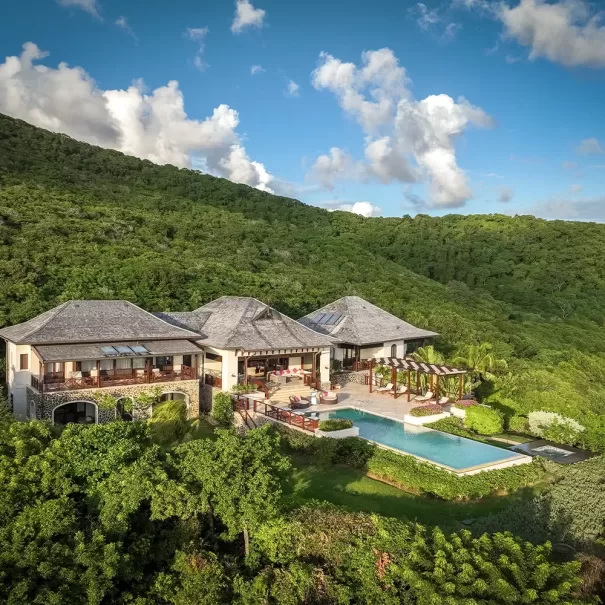 A large luxury villa with a pool sits surrounded by lush green hills under a blue sky with scattered clouds. The house features multiple roofs and balconies, with landscaped gardens and outdoor seating areas.