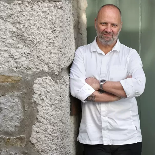 Chef Simon Rogan, wearing a white chefs coat, stands with arms crossed against a stone wall next to a green door, smiling slightly at the camera.