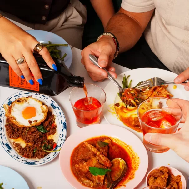 Three people share a colorful meal at a table, serving dishes like curry, a fried egg over minced meat, and salad. One person pours a dark drink into a glass while others toast or eat with forks.
