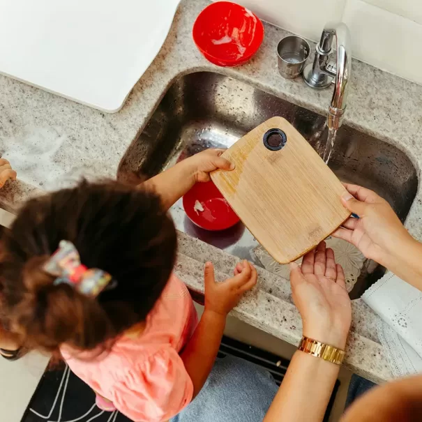A child and an adult are washing a small wooden cutting board in a kitchen sink. The countertop is cluttered with soap, sponges, and a red bowl. The child is wearing a pink shirt, and the adult is wearing a watch on the left wrist.