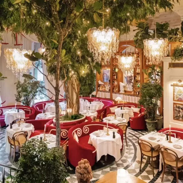 Elegant restaurant with red velvet seating, white tablecloths, lush indoor trees, and large chandeliers. The room features patterned tile floors, greenery, and a warm, inviting atmosphere with soft, ambient lighting.