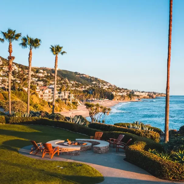 Sunny coastal view with palm trees, a fire pit area surrounded by chairs, manicured grass, and ocean waves. Hills with houses overlook the sandy beach and blue sea under clear skies.