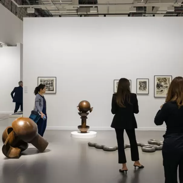 People observe sculptures and framed art on display in a modern gallery. The sculptures are abstract and metallic, and the gallery has bright lighting with a plain white wall as the backdrop.