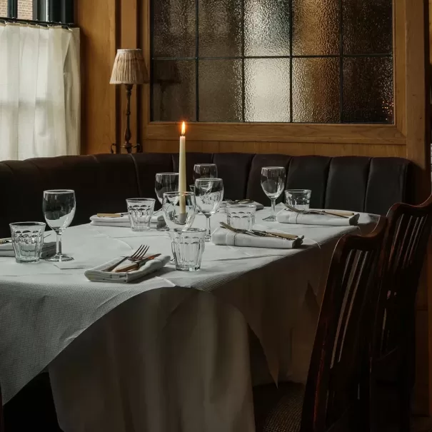 A dimly lit restaurant table set for four with white tablecloth, glassware, plates, cutlery, and a single lit candle, surrounded by dark wooden chairs and a cushioned booth.