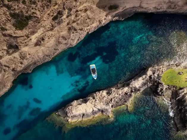 Aerial view of a small boat floating in clear turquoise water between rocky shorelines, with underwater rocks visible and sunlight illuminating the scene.
