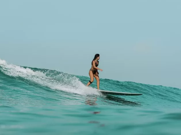 A woman in a swimsuit is surfing on a wave in the ocean. She is standing on a surfboard, riding along a crest with water splashing around. The sky is clear, adding to the serene and adventurous atmosphere.