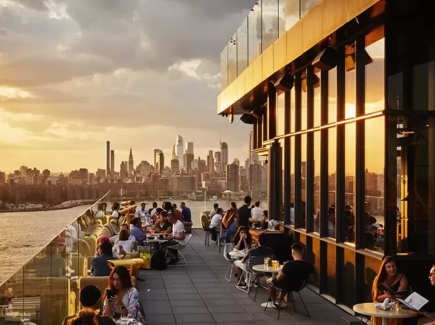 People dining and socializing on a rooftop terrace at sunset, with glass railings overlooking a city skyline full of tall buildings and skyscrapers under a partly cloudy sky.