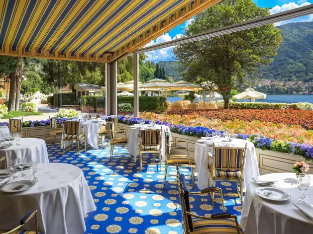 An elegant outdoor restaurant with round tables and striped chairs overlooks a lake and mountains. Bright flowers line the terrace, and a blue patterned carpet covers the floor under a striped canopy.