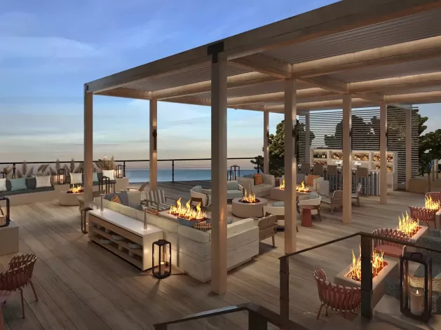 Rooftop lounge at sunset with modern seating, fire pits, wooden pergolas, and ocean views. Cozy sofas and chairs are arranged around low tables, creating an inviting atmosphere for relaxation.