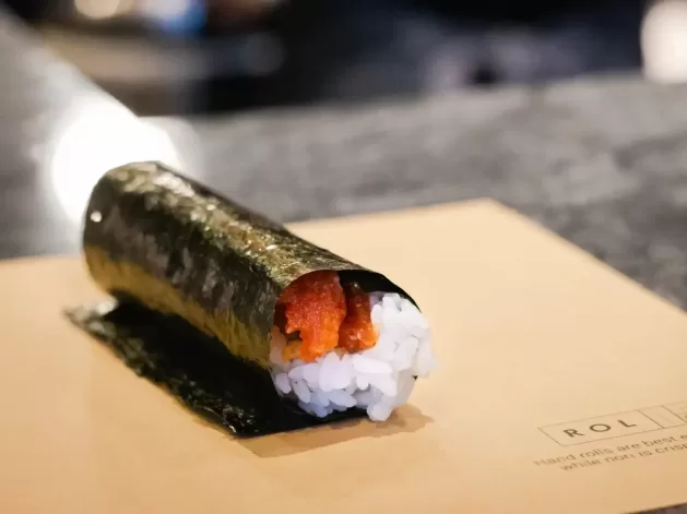 A hand roll sushi with white rice and orange filling, wrapped in a sheet of seaweed, sits on a brown paper surface with blurred background.