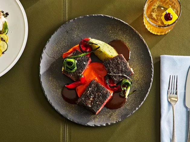 A dish with seared meat, drizzled with red and brown sauce, garnished with roasted red peppers and greens on a black plate. An avocado slice is at the top. Beside it, a glass with an orange liquid and ice, garnished with a cherry, is visible.