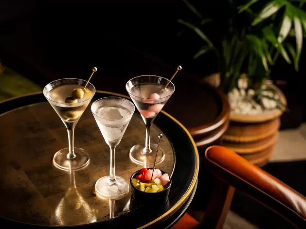 Three martini glasses with olives and cocktail onions are arranged on a round, reflective table next to a small bowl of garnishes. There is an orange chair and a potted plant in the background.