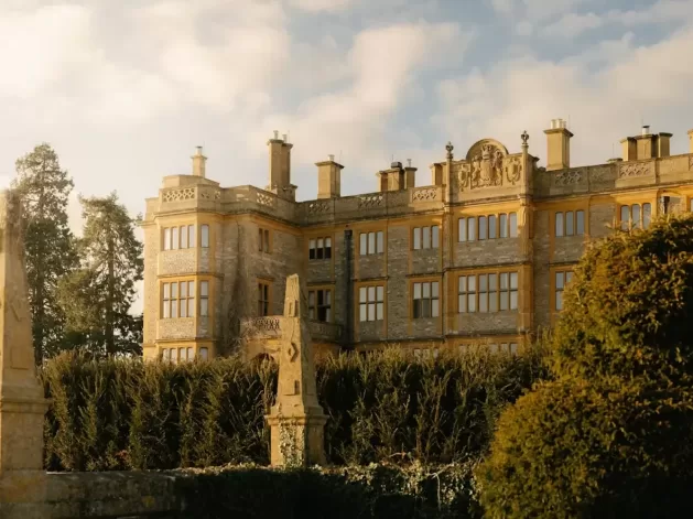 A grand stone mansion with many windows, ornate architectural details, and multiple chimneys stands behind tall hedges on a sunny day, with a lightly clouded sky above.