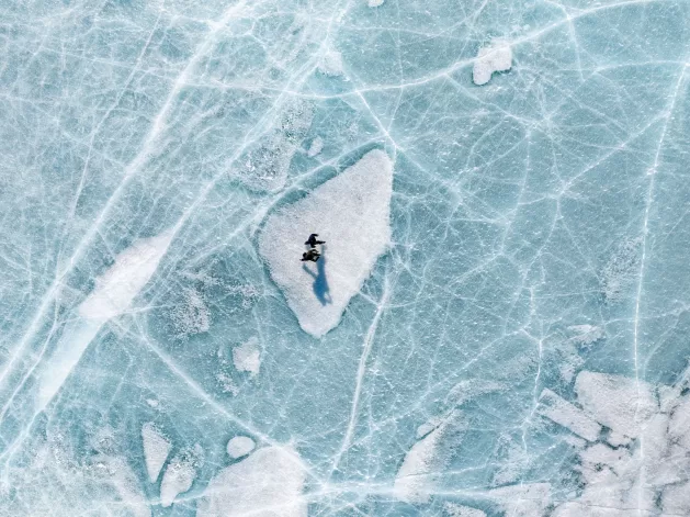 A person stands on a white patch of ice, surrounded by blue cracked and textured frozen lake surface, casting a long shadow. The scene is viewed from above.
