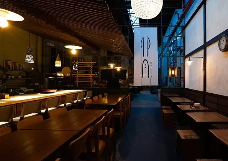 A dimly lit restaurant with wooden tables and chairs. A large, circular hanging light illuminates the space. A white banner with abstract symbols hangs near the back, and theres ambient, warm lighting throughout the interior.