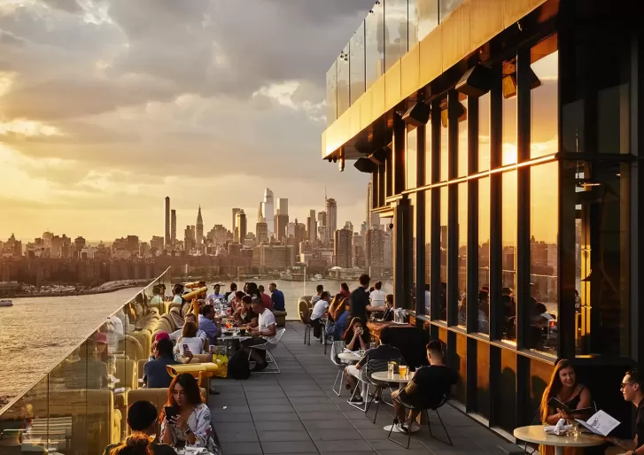 People dining and socializing on a rooftop terrace at sunset, with glass railings overlooking a city skyline full of tall buildings and skyscrapers under a partly cloudy sky.