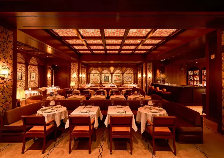 Elegant restaurant interior with warm lighting, featuring rows of tables set with white tablecloths and wooden chairs. The decor includes patterned wallpaper, framed artwork, and an inviting bar area in the background.