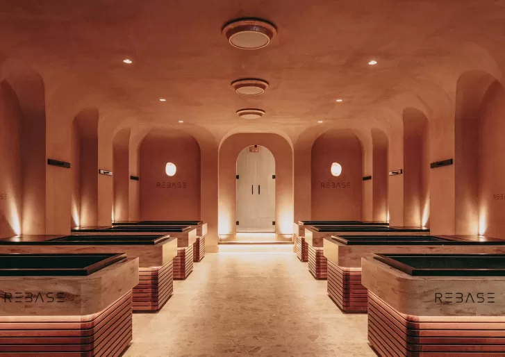 A warmly lit, modern spa room with ten marble soaking tubs arranged in two rows, arched ceilings, and soft ambient lighting creating a calm, inviting atmosphere. The word REBASE is displayed on the tubs and walls.