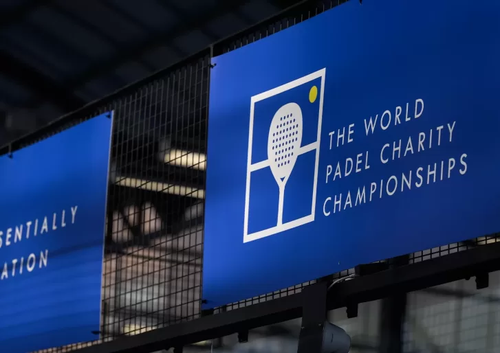 Blue banners hang on a metal fence; one reads QUINTESSENTIALLY FOUNDATION, and the other shows a padel racket graphic with THE WORLD PADEL CHARITY CHAMPIONSHIPS in white text.